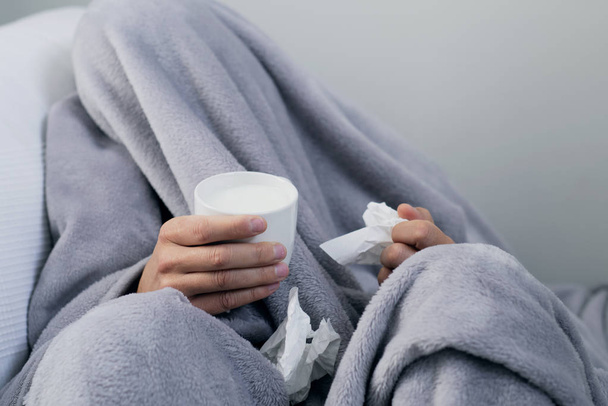 Close-up van een zieke jonge Kaukasische man thuis, bedekt met een licht grijze zachte deken, warming-up met een kop warme mil - Foto, afbeelding