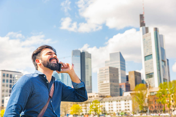 Young man talking on the phone in Frankfurt. Middle eastern man with beard having a call along Main river and laughing. City life and communication concepts - Photo, image