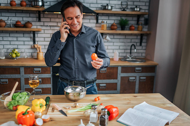 Busy man stand at table in kitchen and talk on phone. He smile and hold egg in hand. Guy cooking. Colorful vegetables on table. - Photo, image