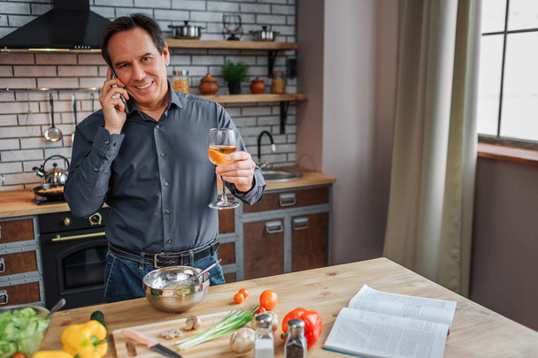 Happy adult man stand at table in kitchen and talk on phone. He hold glass of white wine and smile. Man cooking. Vegetalbes and cookbook on table. - Fotografie, Obrázek