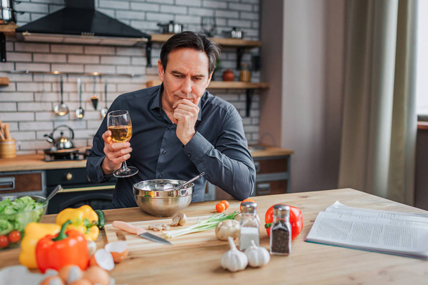Serious man lean to table in kitchen and look down. He hold glass of white wine and hand on chin. Colorful vegetables and spices with journal on table. - Фото, изображение