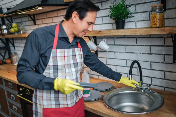 Nice man in apon stand in kitchen and touch sink. He hold plate. Guy wear washing gloves. - Foto, Imagem