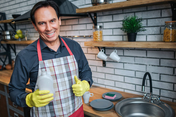 Positive nice adult man stand at sink in kitchen and pose on camera. He hold washing gel and smile. Guy show big thumb up. - 写真・画像