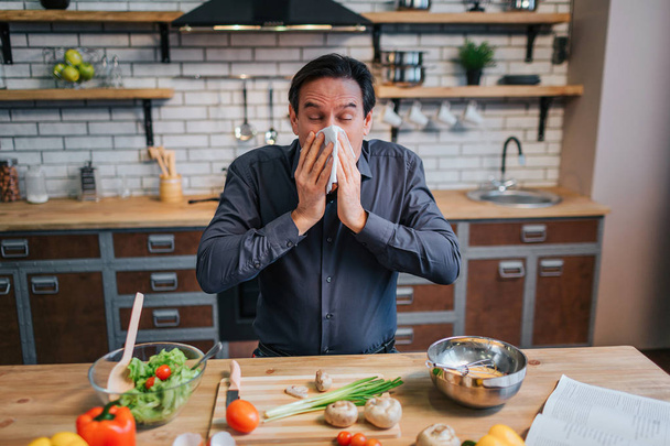 Sick adult man sneezing to white napkin. He stand at table in kitchen. Desk full of colorful healthy vegetables and spices. - Foto, Bild
