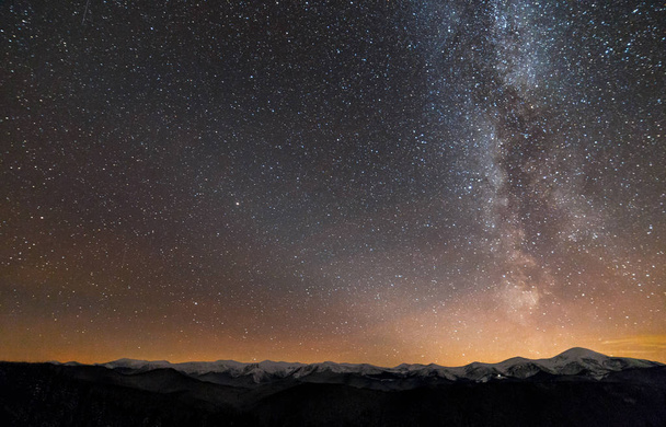 Winter mountains night landscape panorama. Milky Way bright constellation in dark starry sky, soft glow on horizon after sunset, magnificent mountain ridge snow-capped peaks, snowy steep woody hill. - Фото, зображення