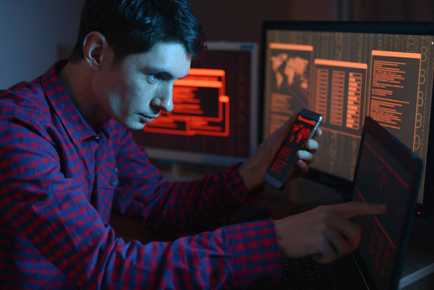 The male hacker in the hood is pointing his finger at the display indicating the location of the cyberattack and hacking data on the screen background in the dark. The concept of cyber security - Zdjęcie, obraz