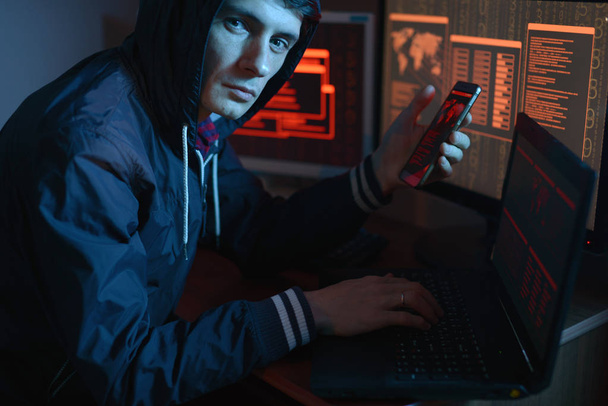 Hombre en el capó mirando a la cámara. Ataques de hackers y fraude en línea en la oscuridad en el fondo de la pantalla en luz de neón. El concepto de ciberseguridad
 - Foto, Imagen