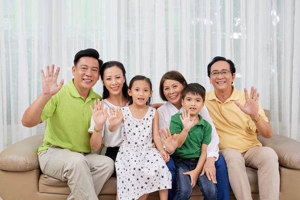 Onnellinen iso aasialainen perhe istuu sohvalla ja vilkuttaa kameralle
 - Valokuva, kuva
