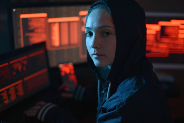 Девушка в капюшоне смотрит в камеру. Хакерские атаки и онлайн-мошенничество на фоне экрана в темноте. Концепция кибербезопасности
 - Фото, изображение