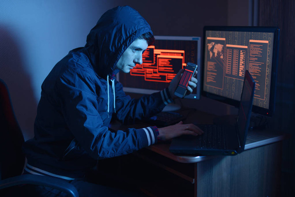 Male hacker in the hood holding the phone in his hands trying to hack the mobile device cloud and steal data in the dark under neon light. The concept of cyber security - Photo, Image