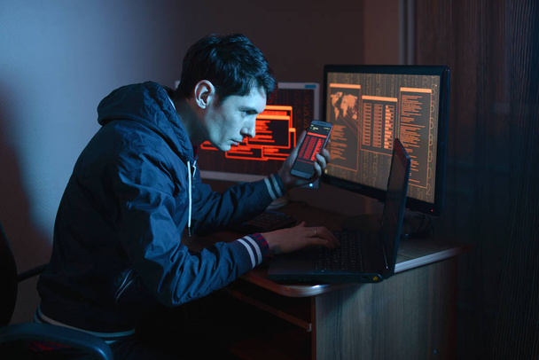 Male hacker holding the phone in his hands trying to hack the mobile device cloud and steal data in the dark under neon light. The concept of cyber security - Photo, Image