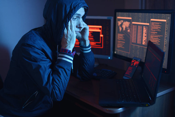 A male hacker in a hood thinking about the problem of hacking or malware infection on the background of screens with codes in neon light. The concept of cybersecurity - Photo, Image