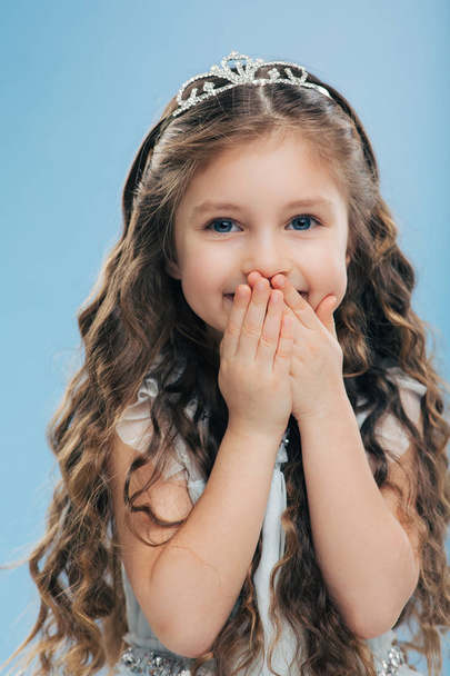 Image of positive beautiful smiling child keeps both hands on mouth, has long thick hair, blue eyes, wears crown, poses against blue background, expresses happiness. Children and satisfaction - Photo, Image
