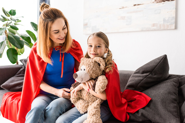 Beautiful mother and kid in red cloaks holding teddy bear and sitting on sofa - Photo, Image