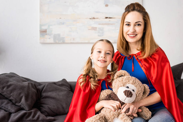 smiling mother and kid in red cloaks holding teddy bear and looking at camera - Photo, Image