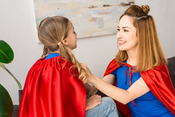 Cheerful mother tying red cloak on daughter at home - Photo, Image
