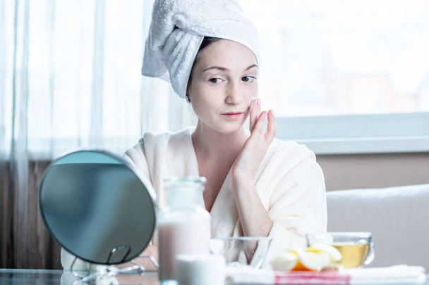 Mulher jovem feliz bonita com uma toalha na cabeça aplicando creme de cosméticos naturais no rosto hidratando a pele. O conceito de higiene e cuidado da pele em casa
 - Foto, Imagem