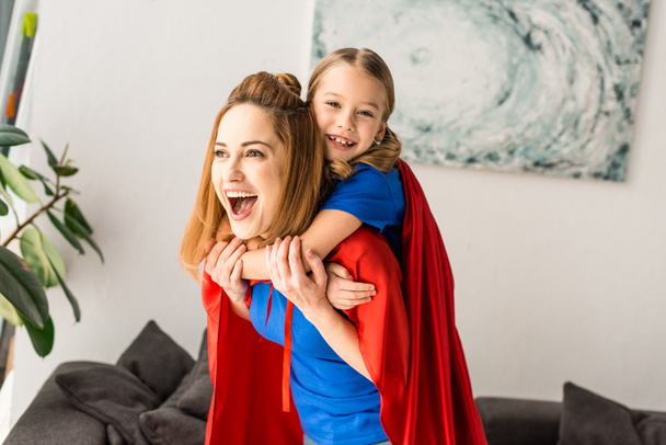 Alegre madre e hija en capas rojas jugando en casa
 - Foto, Imagen