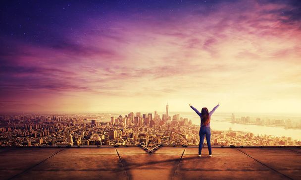 Vista trasera de una joven alegre sentir la libertad levantando las manos de pie en la azotea de un rascacielos viendo la puesta de sol sobre el horizonte de la gran ciudad
. - Foto, imagen