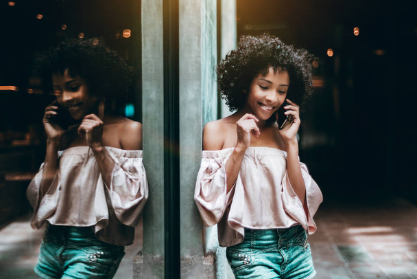 A young flirtatious curly-haird biracial girl is having a phone conversation with her boyfriend and smiling while leaning against the wall with the mirror window in front fully reflecting her - Fotografie, Obrázek