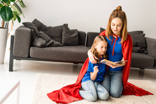 Cute kid and mother in red cloaks sitting on floor and reading book - 写真・画像