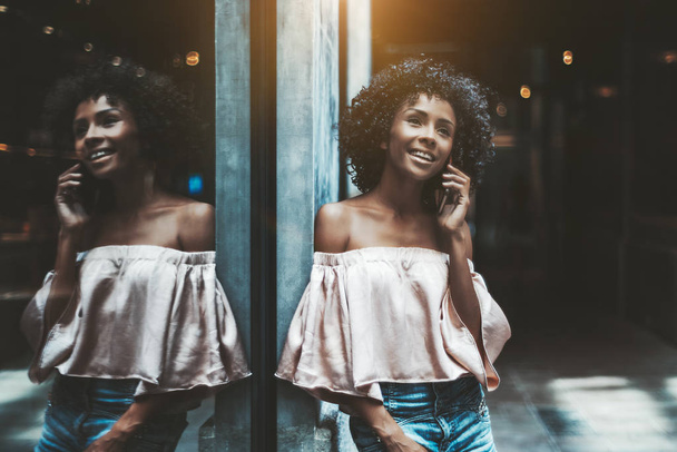A cheerful curly-hair young biracial woman is smiling and talking on the phone with her friend while leaning against the wall with a glass mirror window in front which is fully reflecting her - Photo, image