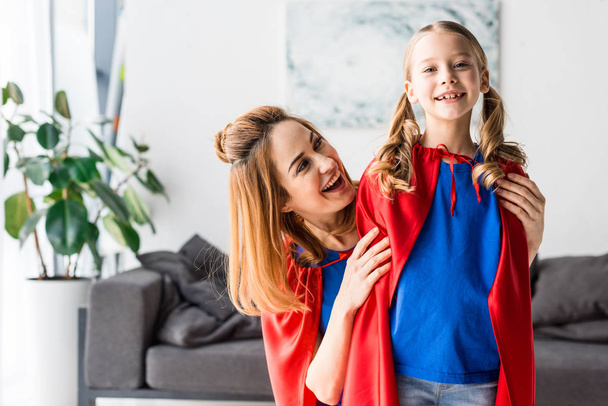 Cute kid and attractive mother in red cloaks smiling and hugging at home - Photo, Image