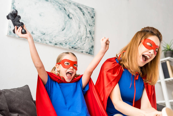 Mother and daughter in red masks and cloaks playing video game at home - Photo, Image