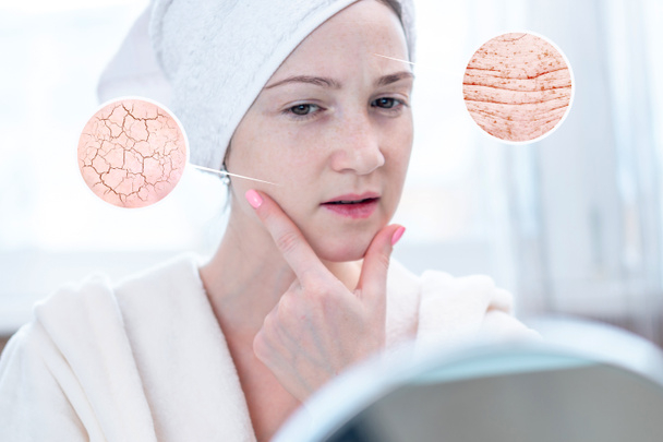 Beautiful young woman with a towel on her head looking at her dry skin with cracks and with first wrinkles. Circles increase the skin like a magnifying magnifier - Photo, Image