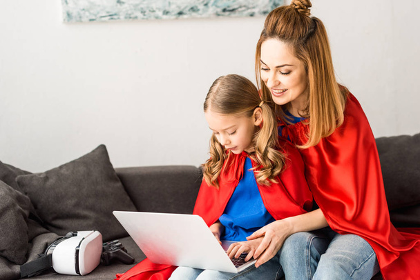 mother and daughter in red cloaks sitting on sofa and typing on laptop - Фото, изображение