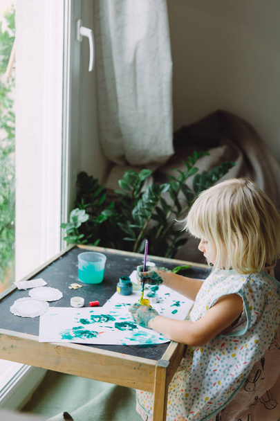Adorável menina brincando com tinta verde em casa perto da janela
 - Foto, Imagem