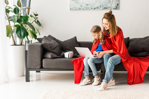 Mutter und Tochter in roten Mänteln sitzen auf Sofa und halten Laptop - Foto, Bild