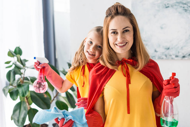 Mother and daughter in red capes and rubber gloves with sprays cleaning and looking at camera - Photo, image
