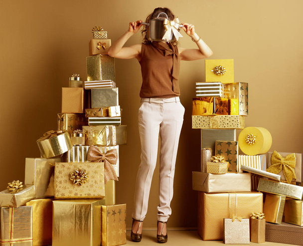 trendy woman in gold beige pants and brown blouse with a stew pan with golden bow as a received bad gift among 2 piles of golden gifts in front of a plain wall. Don't be a bad gift giver concept. - Fotoğraf, Görsel