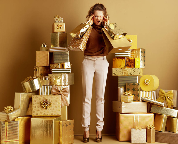 Stressed elegant shopper woman in gold beige pants and brown blouse among 2 piles of golden gifts in front of a plain wall showing shopping bags. where to get money for the holidays concept. - Фото, изображение