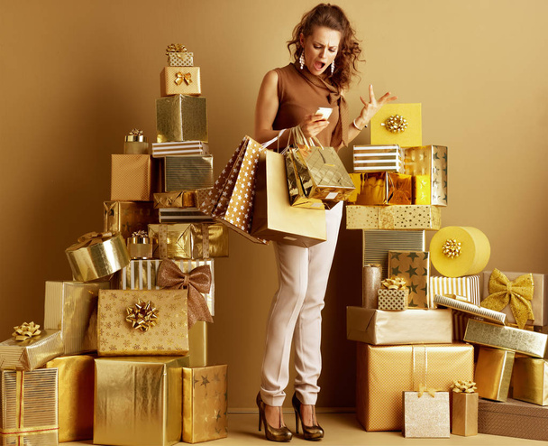 Full length portrait of annoyed modern fashion-monger in gold beige pants and brown blouse with shopping bags looking at smartphone among 2 piles of golden gifts. stressing while planing party concept. - Fotografie, Obrázek