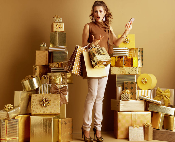 Disappointed stylish shopper woman in gold beige pants and brown blouse with shopping bags and smartphone among 2 piles of golden gifts. cant find a satisfying product from the current supply.   - 写真・画像