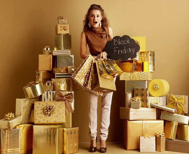 Full length portrait of surprised modern fashion-monger in gold beige pants and brown blouse with shopping bags showing Black Friday sign among 2 piles of golden gifts in front of a plain wall. - Photo, image