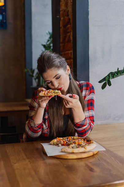 Beautiful and sexy girl sitting by the window in a cafe and eating pizza. - Photo, Image