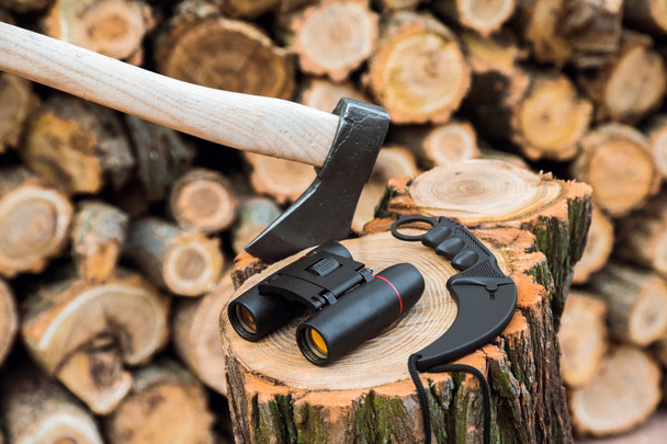 Axe, távcsövek és karambit kés esetén a csonkot, firewoods a háttérben - Fotó, kép