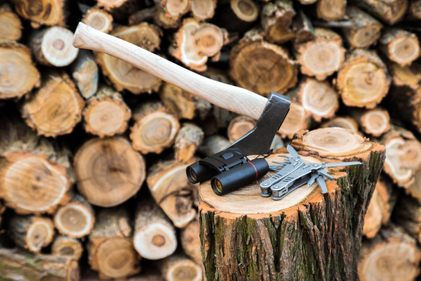 axe, binoculars and multitool knife case on the stump, firewoods on the background - Photo, Image