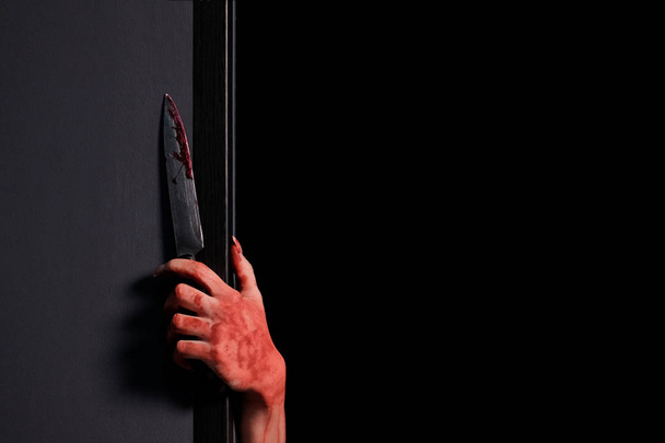 A woman's bloodied hand stuck a knife out from behind a wall.Concept of celebrating Halloween, horror and fear. Close-up. Copy space. Black background. - Photo, Image