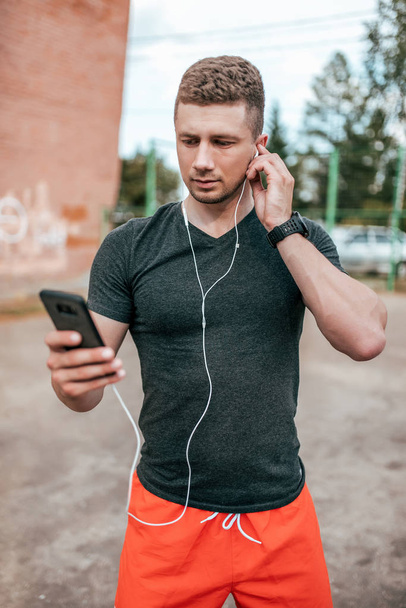 Young guy holding phone listening music with headphones. Summer city sports ground. Checks training on application on Internet. Man on street checks calories eaten. Communication in social networks. - Foto, Imagem