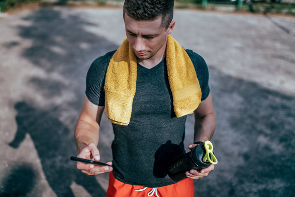 Young guy holding bottle water and phone. In summer in city on sports ground. Checks training on application on Internet. A man with a towel around his neck, on the street checks the calories eaten. - Foto, Imagem