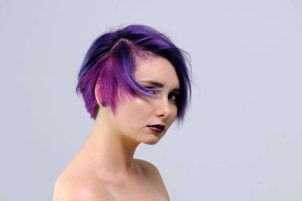 A beautiful, sexy girl with purple hair and a short haircut sits in the middle of the photo with a white background and grimaces. She's naked and visible shoulders. - Foto, imagen