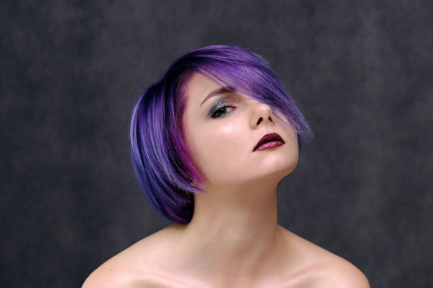 A beautiful, sexy girl with purple hair and a short haircut sits in the middle of the photo with a gray background and grimaces. She's naked and visible shoulders. - Φωτογραφία, εικόνα