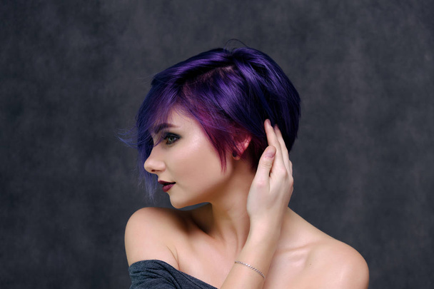 Une belle fille sexy aux cheveux violets et à la coupe courte se trouve au milieu de la photo avec un fond gris et des grimaces. Elle est nue et visible.
. - Photo, image