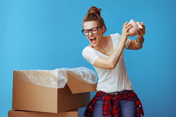 annoyed modern woman in white t-shirt wants to break a piggybank on blue background. Moving can be very stressful and money consuming, but I have to move to a new location. - Foto, Imagen