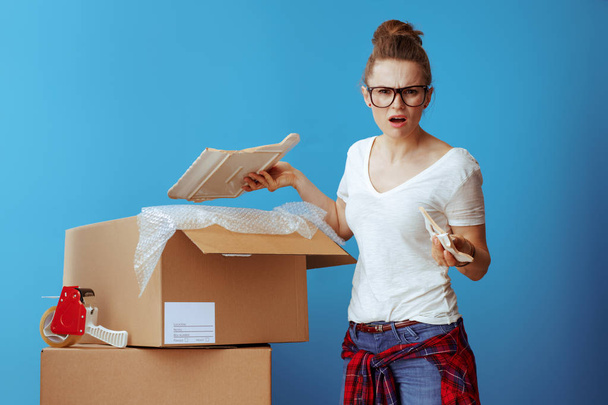 Portrait of unhappy modern woman in white t-shirt near cardboard box with a broken dish on blue background - Fotoğraf, Görsel