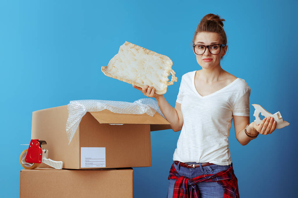 sad modern woman in white t-shirt near cardboard box with a broken dish on blue background - Foto, afbeelding